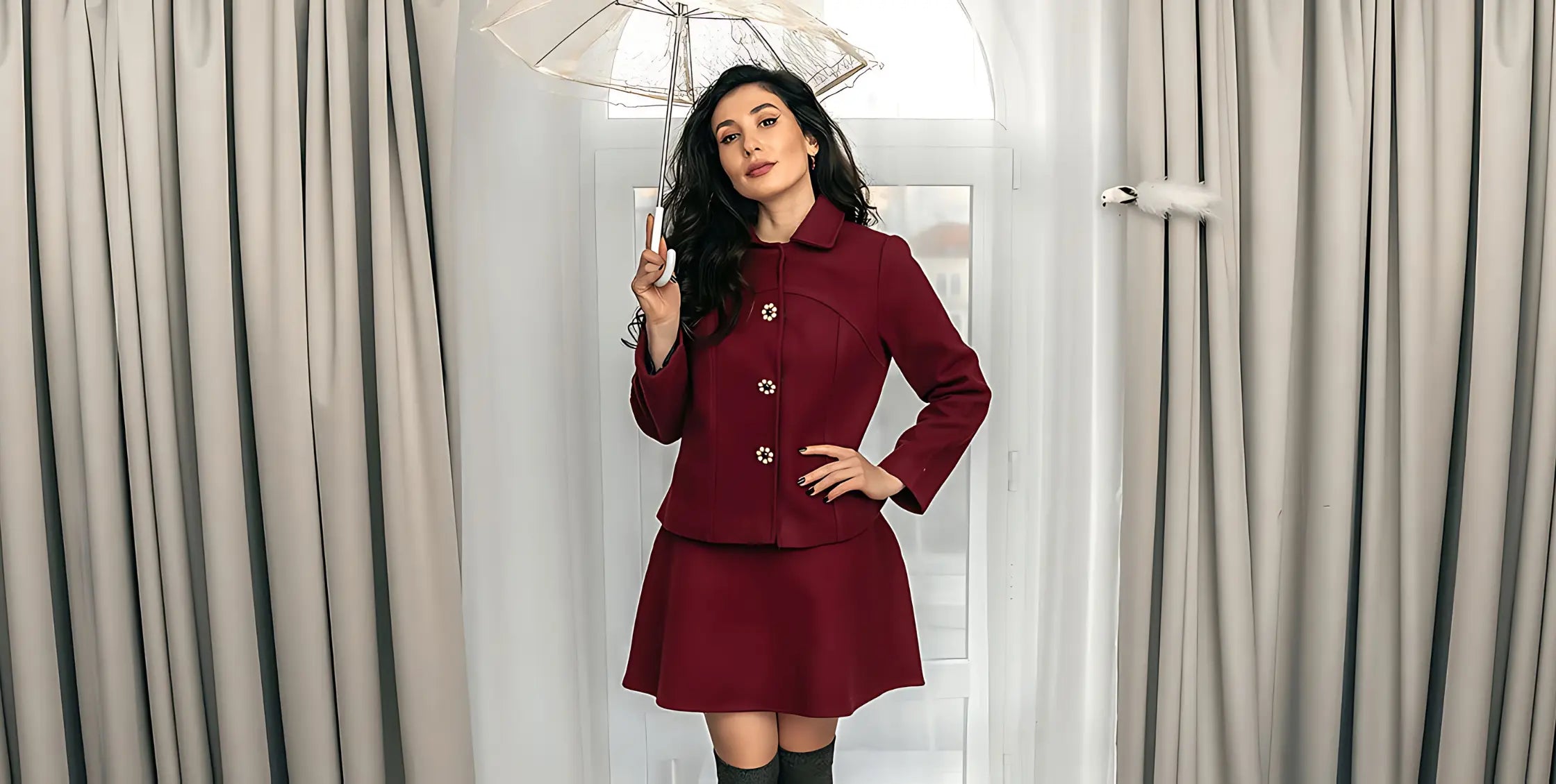Woman wearing a burgundy tailored skirt suit with floral statement buttons, holding a transparent umbrella, standing in front of a white arched window with grey curtains