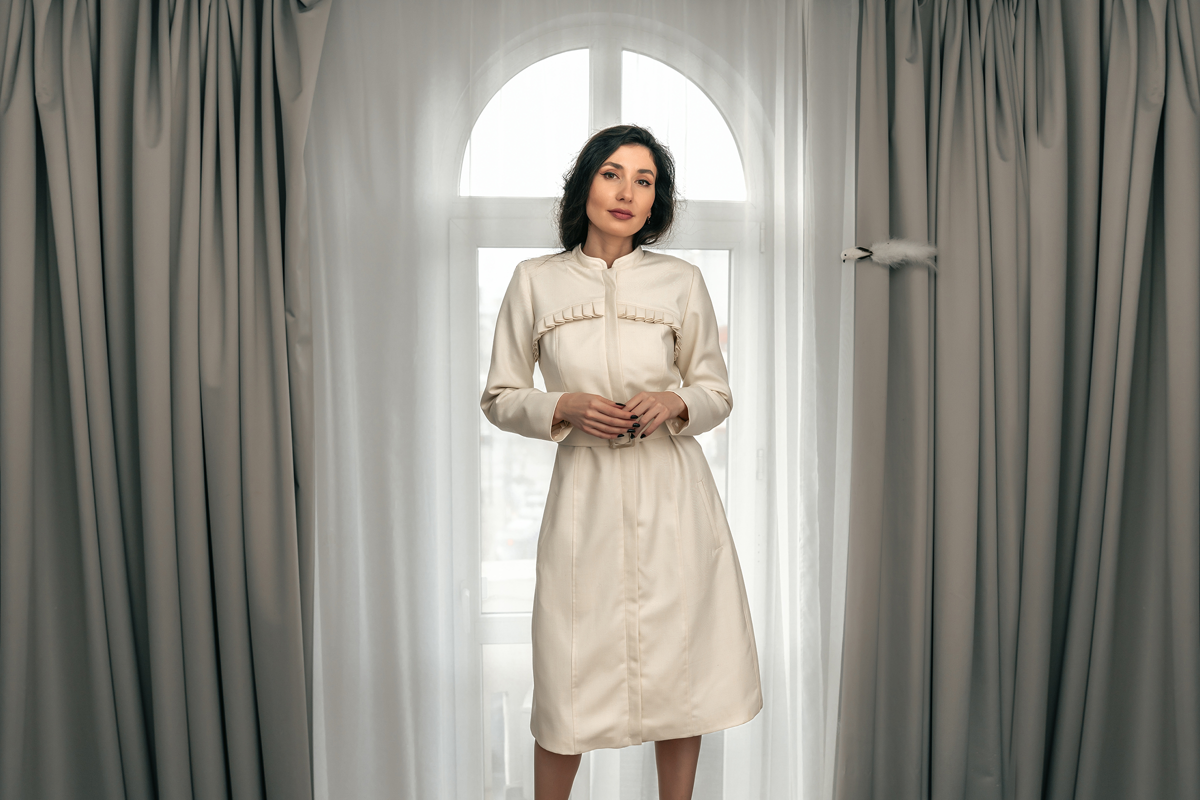 Woman wearing an elegant cream long-sleeve midi dress with a high neck and tailored fit, standing in front of a tall arched window with grey curtains