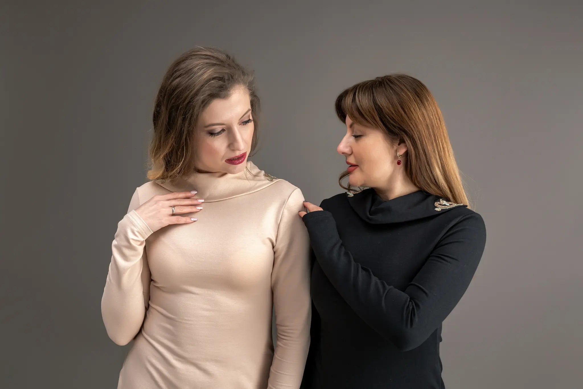 Two women wearing elegant long-sleeve tops, one in light beige and one in black, both with decorative shoulder details, standing against a neutral grey studio background