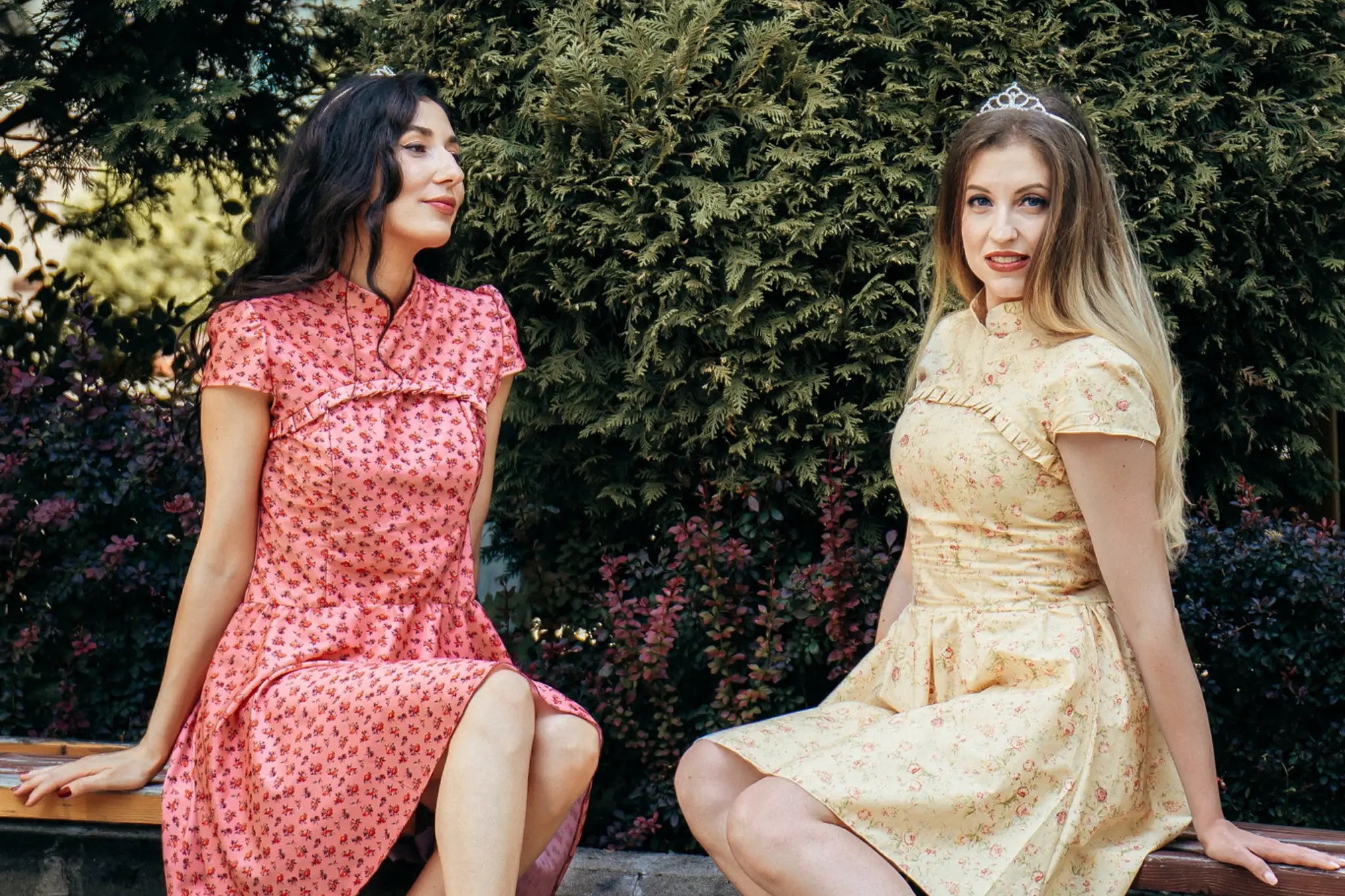 Two women sitting outdoors wearing floral vintage-style dresses, one in pink and one in yellow, featuring short sleeves and fitted waists, with greenery in the background