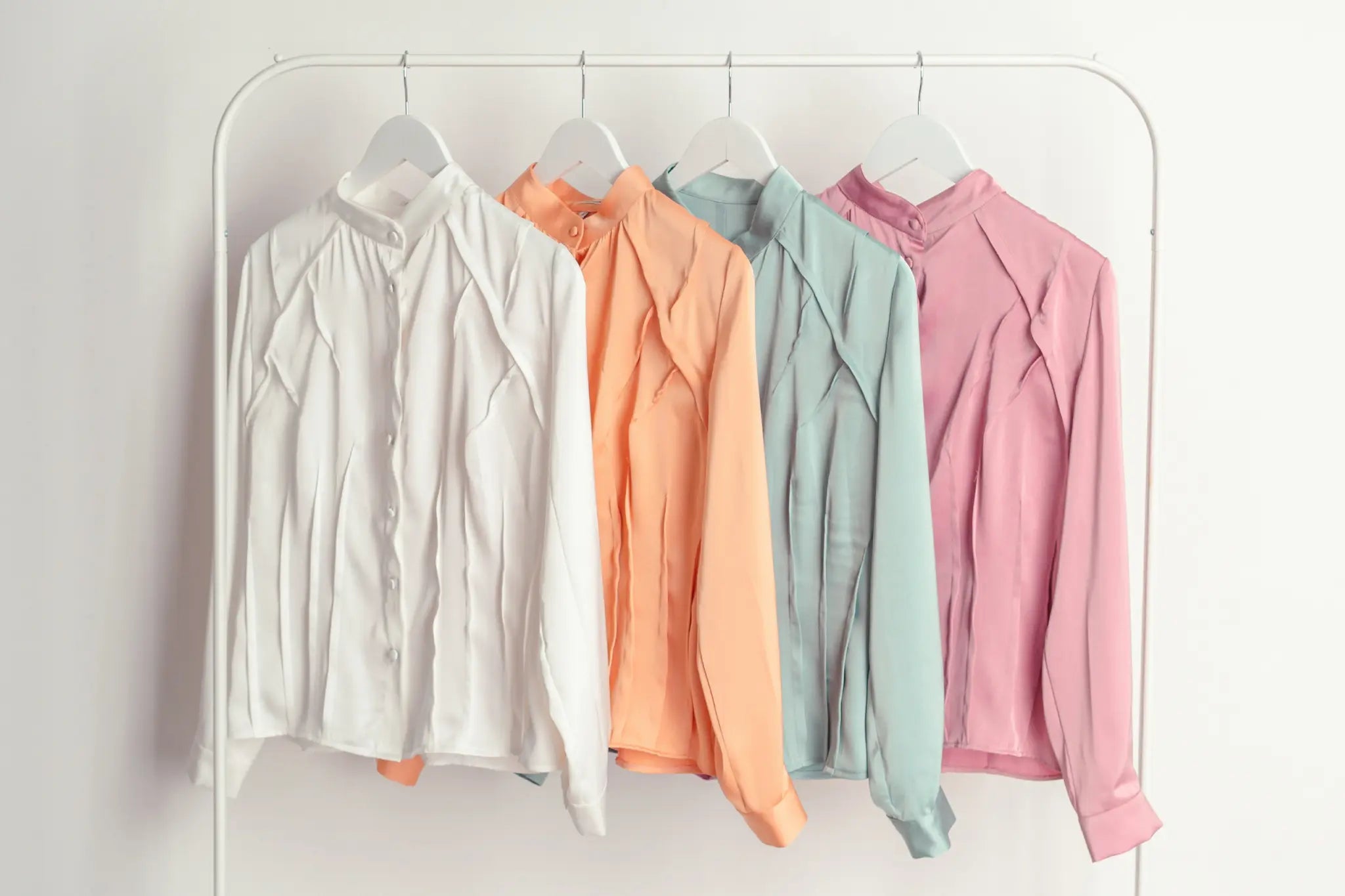 Four satin blouses in white, peach, mint green and soft pink hanging on white hangers from a minimalist clothing rack against a white background