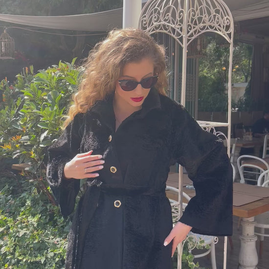 Model wearing a black textured coat with wide sleeves, gold buttons and a belted waist, walking confidently in black heels and sunglasses outside a garden café