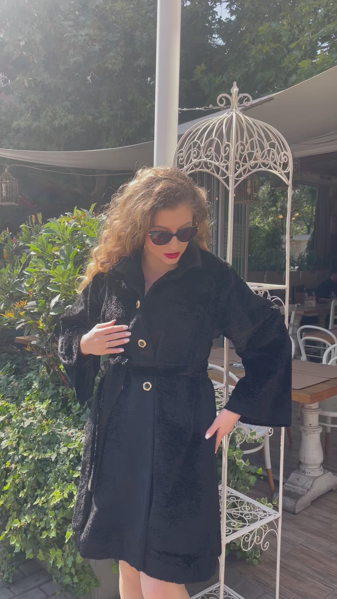 Model wearing a black textured coat with wide sleeves, gold buttons and a belted waist, walking confidently in black heels and sunglasses outside a garden café